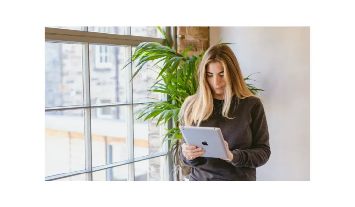 An image of a woman using an iPad standing by a large industrial window with a large green plant behind them.