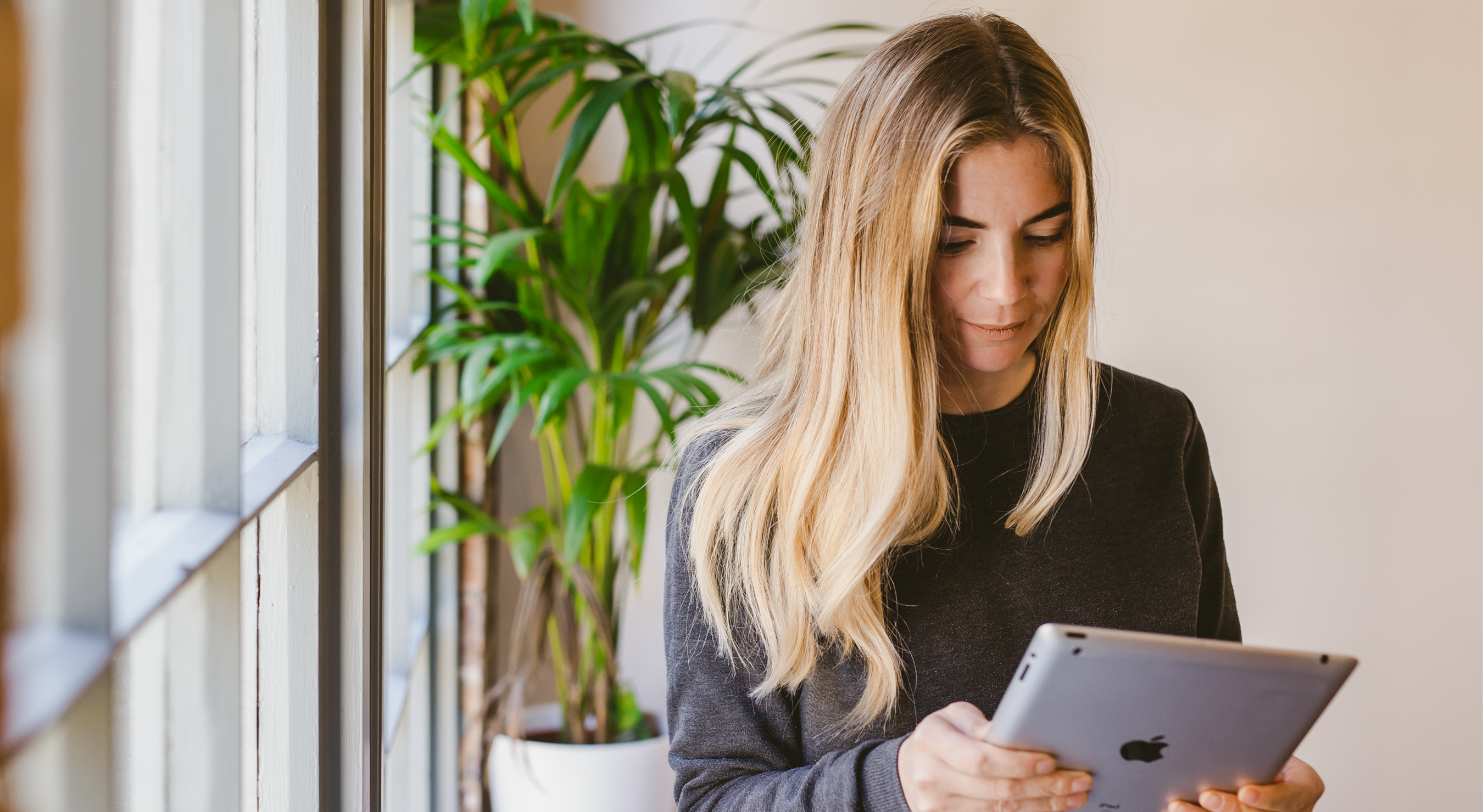 An image of a woman using an identity verification service with her IPad, standing next to a window with a large green indoor plant behind her.