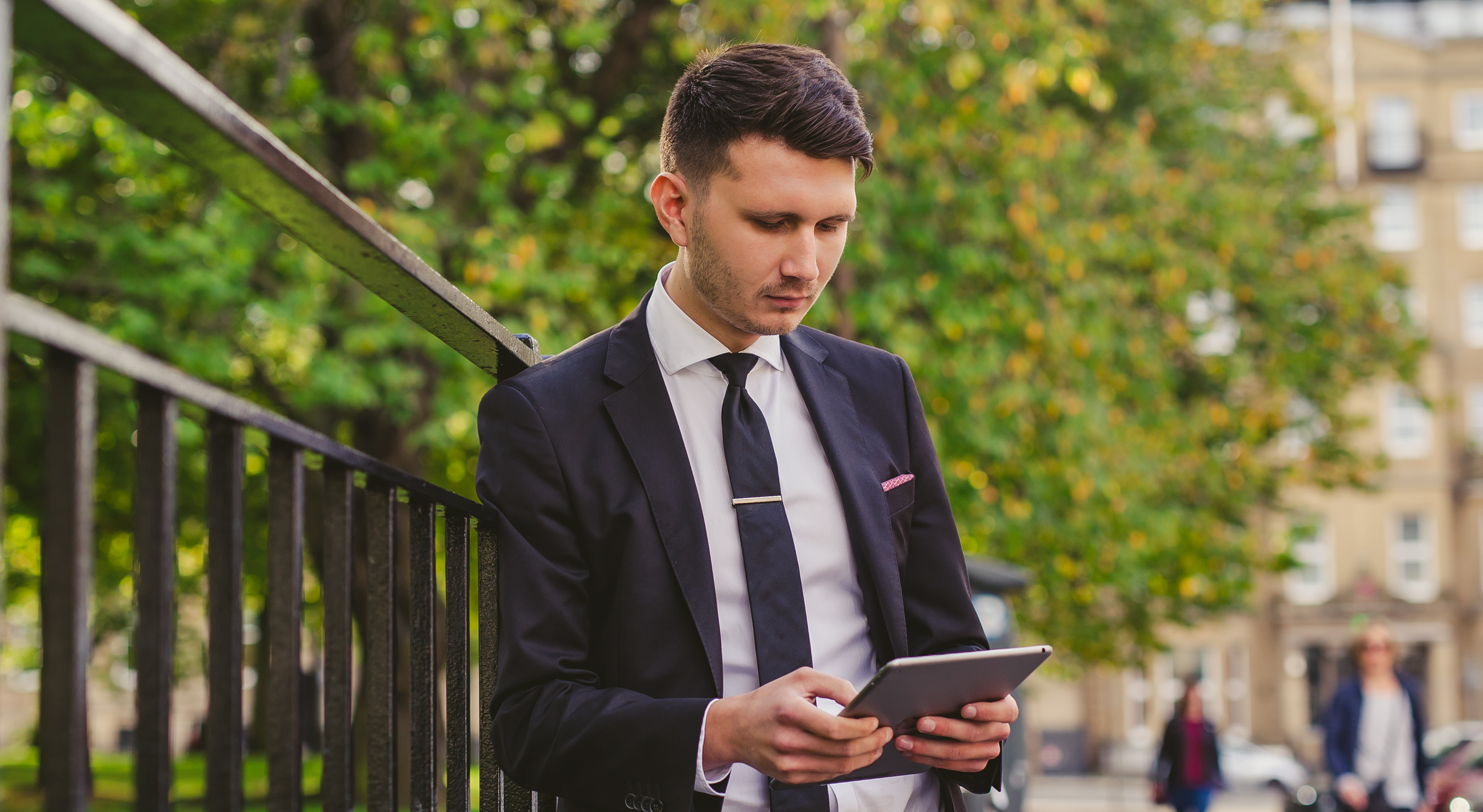 An image of a man holding iPad doing anti money laundering checks in public leaning on a fence with trees behind him.