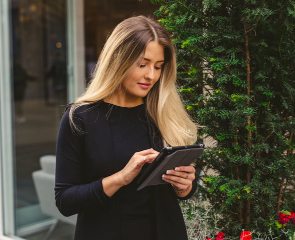 An image of a woman working on a tablet outside standing next to a potted tree.