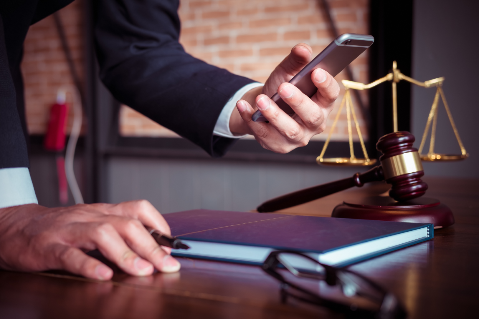 A person wearing a suit holding their mobile phone looking at the screen next to a formal looking book and a gavel in the background.