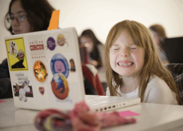 Photograph of a young girl grinning in front of an open laptop. The laptop is covered in stickers. 